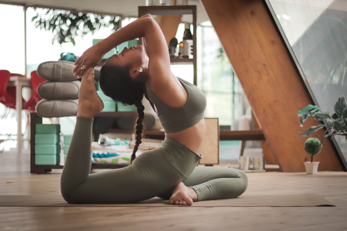 A practitioner in pigeon pose wearing sage green, at home on a woven mat